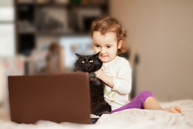 Weinig leuke meisjeszitting op het bed met favoriete huisdierenkat en het gebruiken van een digitaal tabletlaptop notitieboekje. Bel vrienden of ouders online.