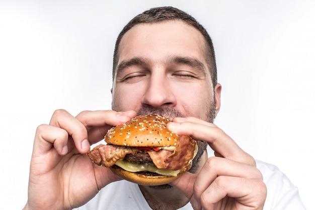 Weird and bizarre man is eating fat and juicy hamburger. It is not a healthy food but the guy likes it very much. His face is very emotional. Isolated on white .