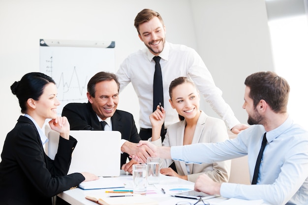 Welcome on board! Group of confident business people in formalwear sitting at the table together and smiling while two men handshaking