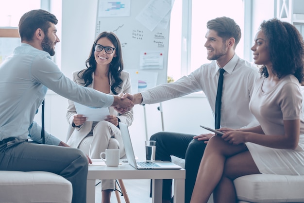 Welcome on board! Two handsome men shaking hands with smile while sitting on the couch at office with their coworkers