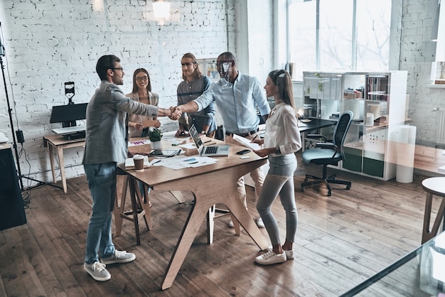 Welcome! Full length top view of young modern men in smart casual wear shaking hands while standing with their colleagues behind the glass wall in the board room