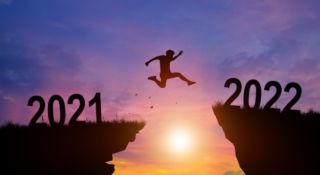 Welcome merry Christmas and Happy new year in 2022. Man jumping across the gap from 2021 to 2022 cliff with Sunset and Twilight Sky background.