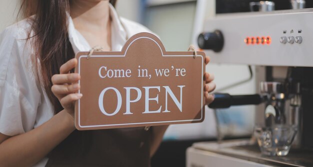 Welcome Open 바리스타 웨이트리스 여성이 카페 레스토랑 소매점 소기업 소유주 음식 및 음료 개념을 서비스할 준비가 된 현대적인 카페 커피숍의 유리문에 열린 간판을 돌리고 있습니다.