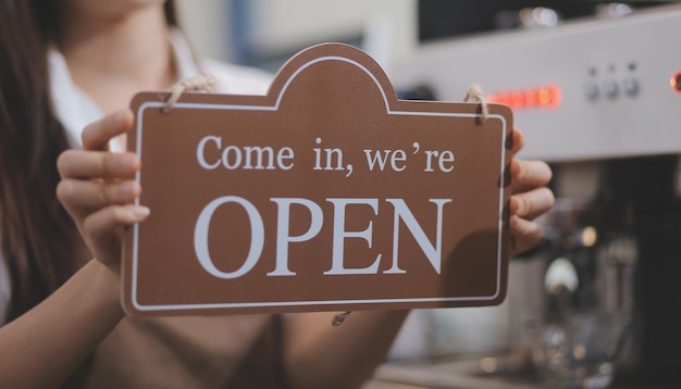 Welcome Open 바리스타 웨이트리스 여성이 카페 레스토랑 소매점 소기업 소유주 음식 및 음료 개념을 서비스할 준비가 된 현대적인 카페 커피숍의 유리문에 열린 간판을 돌리고 있습니다.