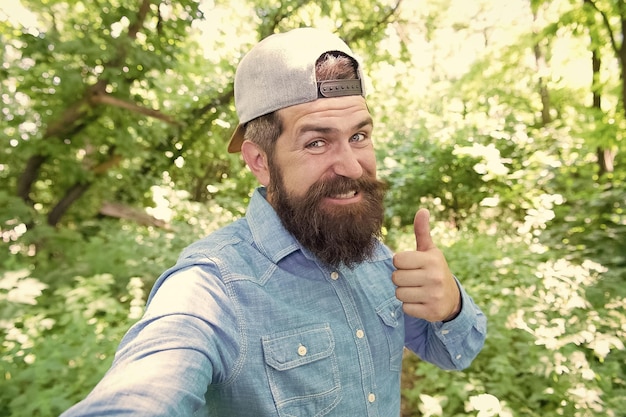 Welcome To Our Campfire mature guy in hat happy hipster traveler making selfie in forest outdoor with sunset nature on background Bearded man casual style fashion portrait of bearded man