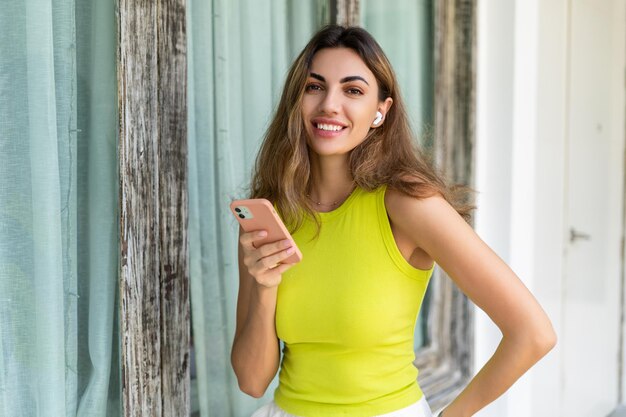 Добро пожаловать Портрет веселой женщины, стоящей на современной вилле со смартфоном и наушниками, держащей телефон со счастливой задумчивой улыбкой