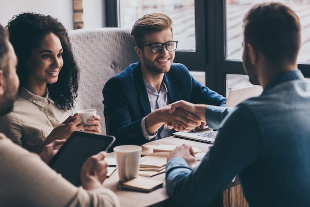 Welcome to team! Two men shaking hands and looking at each other with smile while their coworkers sitting at the business meeting