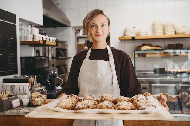 Фото Приветствующая женщина-пекарь держит свежеиспеченные миндальные круассаны на фоне пекарни