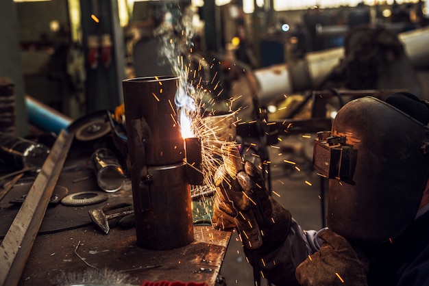 Welder in protective uniform is welding metal pipe in his workshop while sparks flying in front of him.