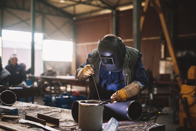 Saldatore in uniforme protettiva e maschera saldatura tubo metallico sul tavolo industriale mentre volano scintille.