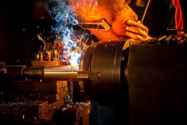 Saldatori in indumenti protettivi e tubi metallici saldano maschere su scrivanie industriali mentre le scintille volano. primo piano di trapani e rottami nei negozi industriali