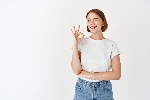 Well done. Smiling positive girl winking, showing okay sign, approve and like something good, praise perfect quality, standing against white wall
