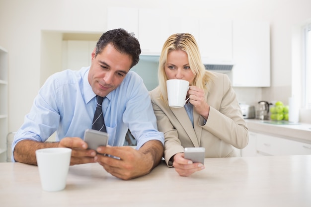 Well dressed couple with coffee cups text messaging in the kitchen