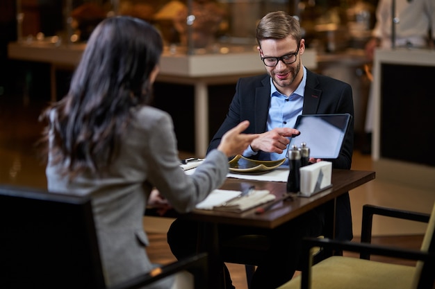 Well-dressed man tilting head down while showing tablet display with finger to a speaking businesswoman before him