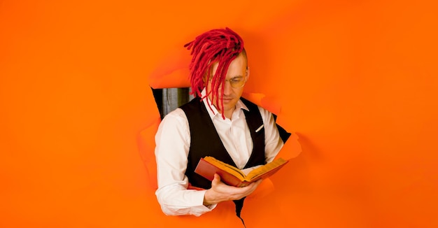 Well dressed man with bright hairstyle reading book and\
sticking out of hole of orange background impressive hipster in\
eyeglasses with red dreadlocks reading old book