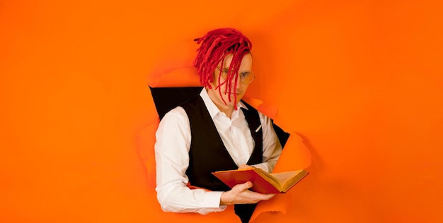 Well dressed man with bright hairstyle reading book and sticking out of hole of orange background Impressive hipster in eyeglasses with red dreadlocks reading old book