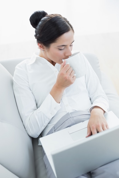 Well dressed woman drinking coffee while using laptop and coffee cup at home