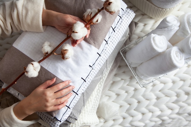 Well groomed woman hand holding a cotton branch with stack of neatly folded linens near rolled up towels in mesh basket placed on knitted chunky merino wool yarn plaid. Natural textile. Top view.