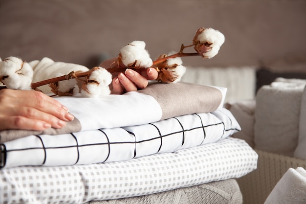 Well groomed woman hands holding the cotton branch with pile of neatly folded bed sheets, blankets and towels. Production of natural textile fibers. Manufacture. Organic product.