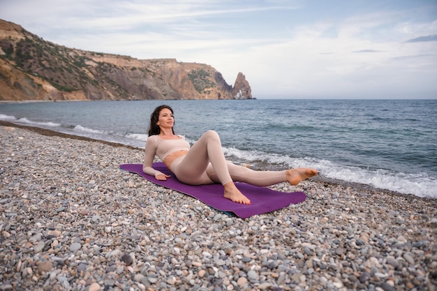 Well looking middle aged woman with black hair fitness instructor in leggings and tops doing stretching and pilates on yoga mat near the sea Female fitness yoga routine concept Healthy lifestyle