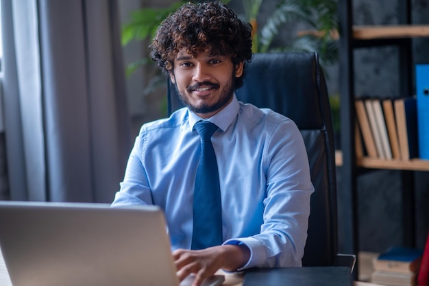 Wellbeing. Curly haired young adult indian man in tie smiling sitting at table working on laptop looking at camera in office
