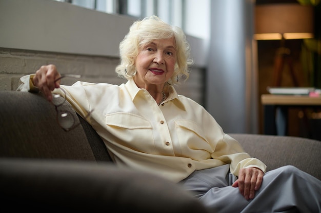 Wellbeing. Mature gray-haired good looking woman holding glasses smiling at camera sitting on sofa near window in cozy room