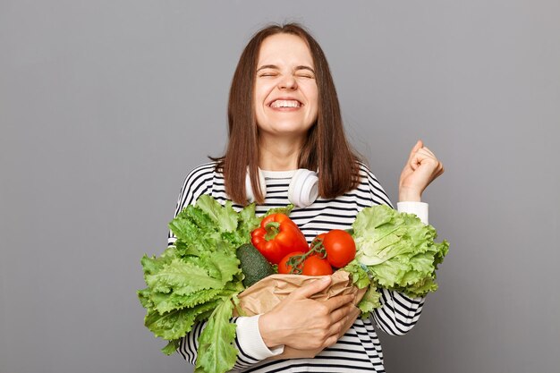 健康は健康的な食事から始まります健康的な食べ物で体に栄養を与えます灰色の背景の上に野菜を保持している幸せな大喜びの女性立っている握りこぶしを祝って立っています