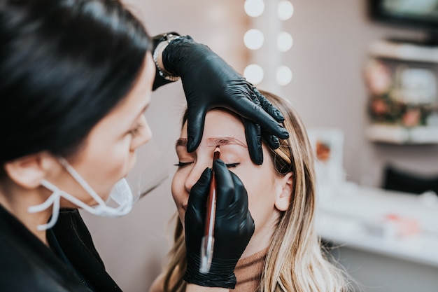 Wenkbrauwen microblading concept, wenkbrauw permanente make-up procedure. Schoonheidsexpert draagt een afgeschermd en beschermend gezichtsmasker vanwege de pandemie van het Coronavirus.