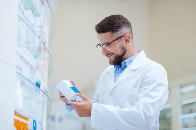 Werk met plezier. Knappe brunette man kijkt naar pakket terwijl hij in de apotheek werkt