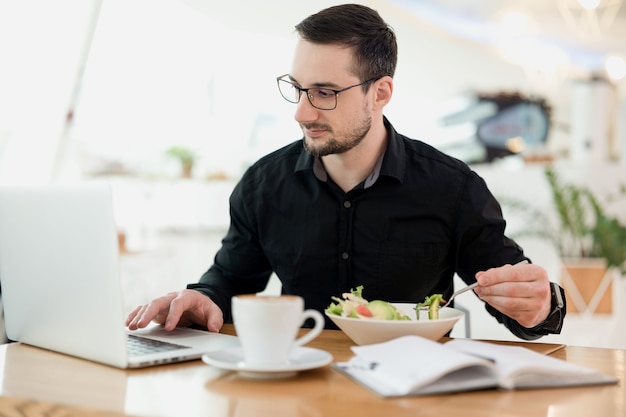 Werk niet tijdens het eten! Geconcentreerde man met baard en bril die geniet van verse salade en iets typt op zijn laptop. Werken op afstand vanuit koffiehuis. Lekkere cappuccino op houten tafel.