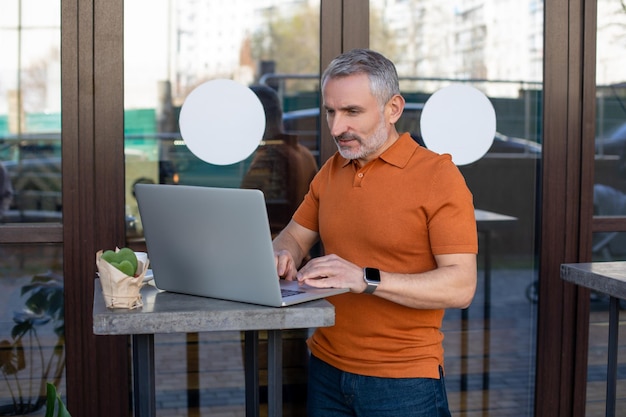 Werk online. Volwassen man in oranje shirt die online werkt in het straatcafé