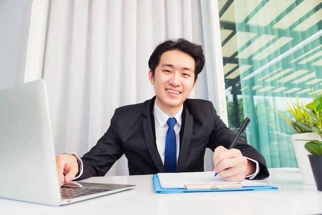 Werk vanuit huis, Aziatische jonge zakenman glimlach draagt pak videoconferentiegesprek of facetime door laptopcomputer op bureau en schrijft notities maken op papier thuiskantoor