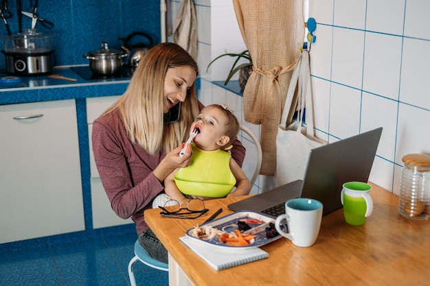 Werk vanuit huis jonge moeder met babymeisje die thuis werkt met behulp van laptop op keukenachtergrond young
