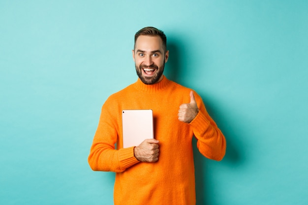 Werk vanuit huis, technologieconcept. Knappe man met laptop, duim opdagen, goedkeuren en iets leuk vinden, staande op turkooizen achtergrond.