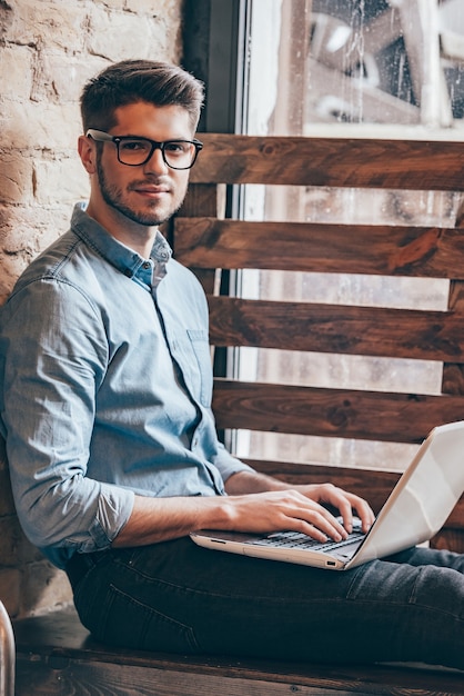 Werken aan zijn blog. Knappe jonge man die op laptop werkt en naar de camera kijkt terwijl hij op de vensterbank zit