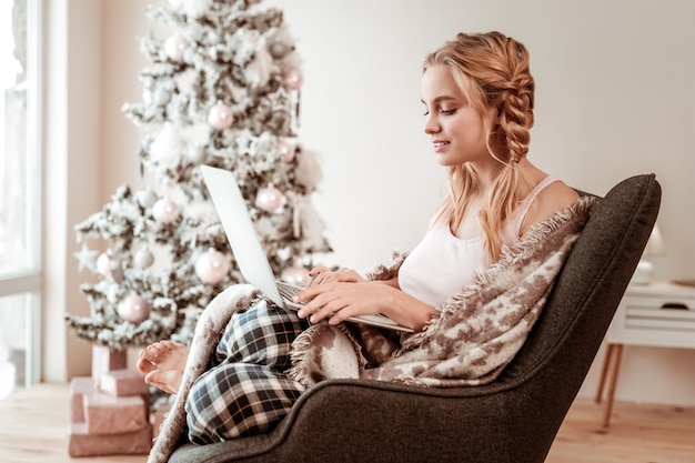 Werken als freelancer. Geconcentreerde langharige vrouw bedekt met deken tijdens het typen op laptop toetsenbord en met kerstboom achter