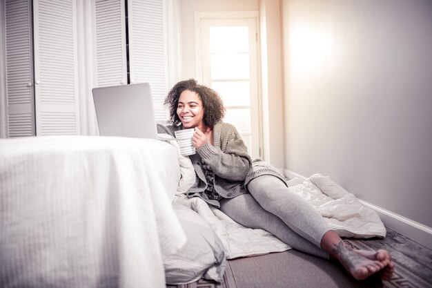Werken met laptop. Vrolijke Afro-Amerikaanse dame zittend op een deken in haar slaapkamer en het controleren van informatie op laptop
