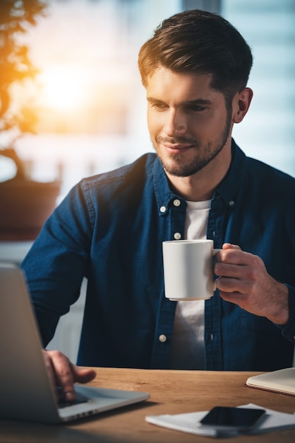 werken met plezier. vrolijke jonge knappe man die een koffiekopje vasthoudt en zijn laptop met een glimlach gebruikt