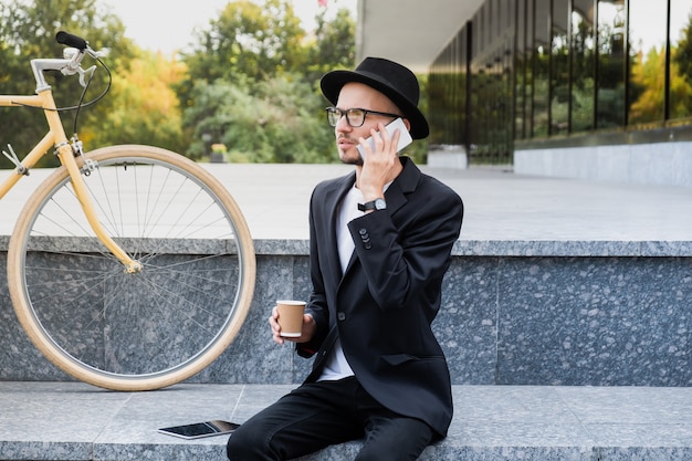 Werken met technologie buiten kantoor. Jonge man in stijlvol casual pak praten aan de telefoon in stedelijk gebied, zittend naast een woon-fiets