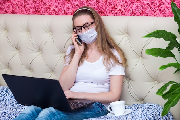 Foto werken op afstand in quarantaine thuis. een mooie blanke meisje werkt op een laptop in een beschermend medisch masker, praten over de telefoon