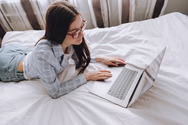 Werken op afstand thuis, vrouw liggend op het bed en werken met een laptop, kijkend op het scherm.