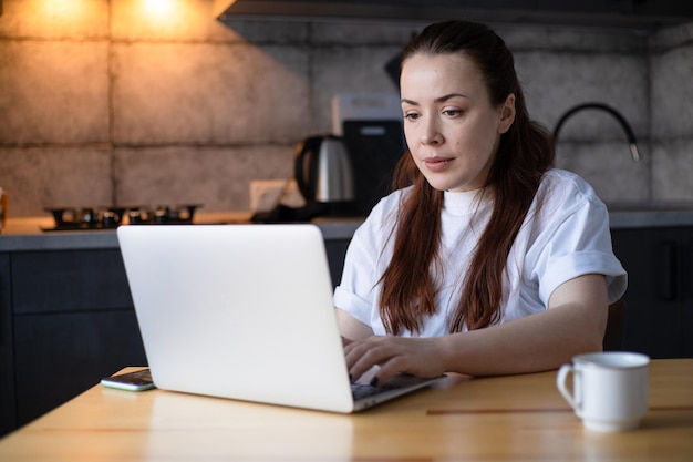 Werken op het keukenkantoor Werken op afstand vanuit kantoor aan huis Jonge vrouw met behulp van laptop Freelancer werkplek aan keukentafel Afstandsonderwijs