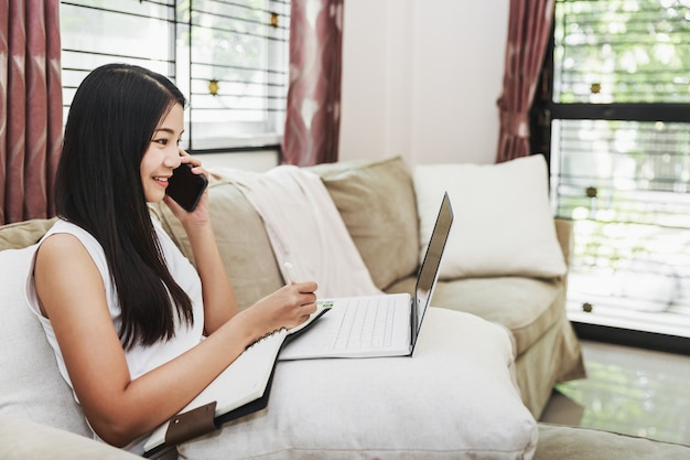 Werken vanuit huis en online winkelen concept, gelukkige aziatische bedrijfsvrouw met behulp van mobiele telefoon en laptopcomputer met laptop op de bank in de huiskamer