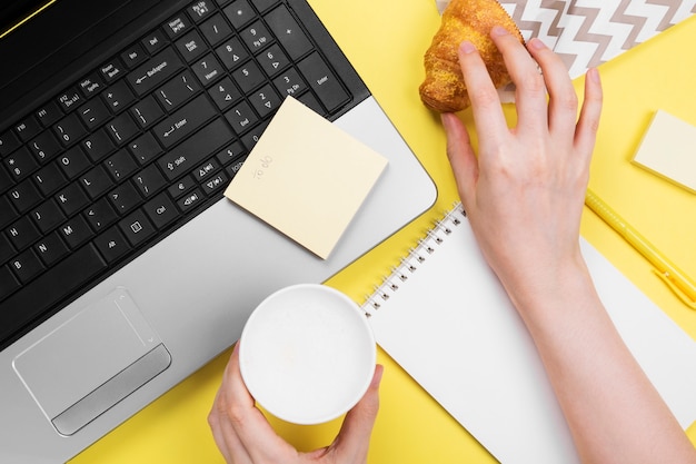 Werkende ochtend. Meisje werkt voor laptop. Ontbijt op het werk - cappuccino, croissant, notebook op gele achtergrond. Bovenaanzicht