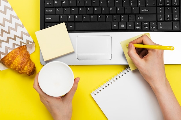 Werkende ochtend. Meisje werkt voor laptop. Ontbijt op het werk - cappuccino, croissant, notebook op gele ruimte. Bovenaanzicht