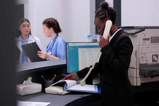 Werknemer die aan de vaste telefoon praat met een patiënt op afstand terwijl hij medische afspraakpapieren controleert, staande aan de balie van de ziekenhuisreceptie. Gezondheidszorgondersteunende service en concept