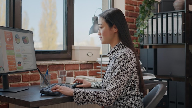 Werknemer die computer gebruikt om aan bedrijfsproject in opstartbureau te werken. vrouw die marketingstrategie plant met gegevensgrafieken op monitor, die helpt bij de ontwikkeling en groei van het bedrijf.