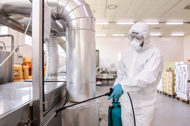 Werknemer in beschermende witte beschermende uniform met masker en rubberen handschoenen sproeier met desinfectiemiddel en spuitmachine in voedselfabriek te houden.