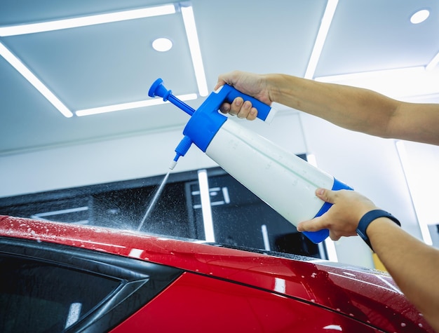 Werknemer in de autoservice spuit water op de auto voordat hij een beschermende film aanbrengt