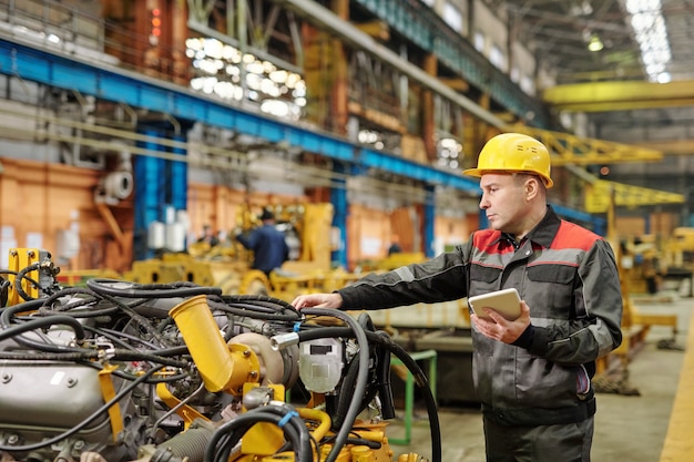 Werknemer in helm die tablet-pc gebruikt en machineapparatuur in de fabriek onderzoekt